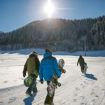 Three snowboarders walk on a snowy slope in bright winter sunlight, heading towards a scenic ski area.